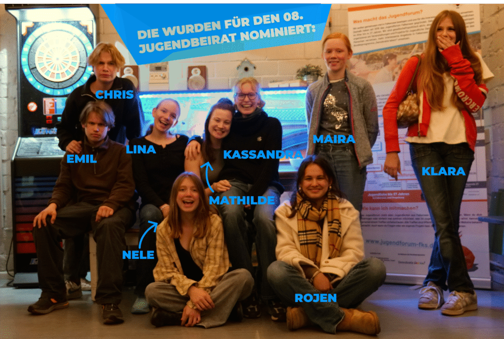 Gruppe von Jugendlichen, die für den 8. Jugendbeirat nominiert wurden, lächeln zusammen. Im Hintergrund ein Dartautomat und ein Informationsplakat.