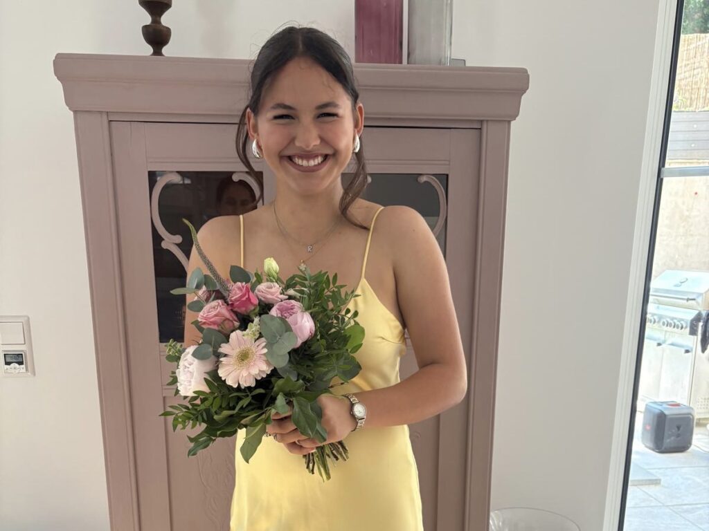 Junge Frau in einem gelben Kleid hält einen bunten Blumenstrauß mit Rosen und Gerbera. Sie lächelt fröhlich vor einem hellen Hintergrund.