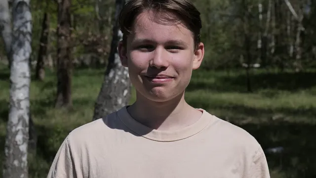 Lächelnder junger Mann mit kurzem, braunem Haar, trägt ein hellbeiges T-Shirt, steht vor einer grünen Waldlandschaft.